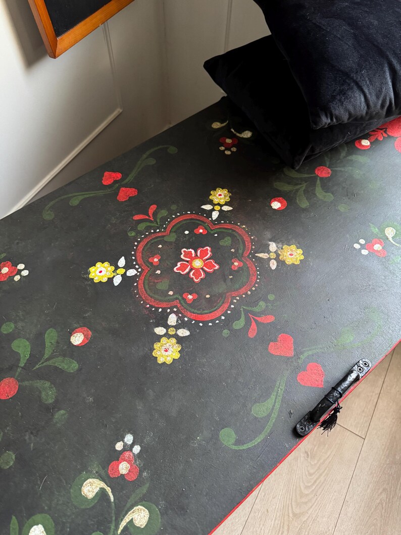 May include: A black wooden storage chest featuring a hand-painted floral motif in red, yellow, and green. The design includes stylized flowers, leaves, and heart shapes across the top surface, which also has a black metal handle attached to the side.