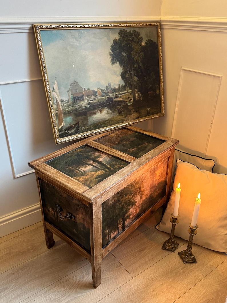 Peut inclure: Un coffre en bois avec une peinture de paysage sur le couvercle et le devant. Au-dessus, une peinture de paysage encadr&eacute;e est accroch&eacute;e au mur. Deux bougies allum&eacute;es dans des bougeoirs en laiton sont plac&eacute;es pr&egrave;s d'un coussin beige.