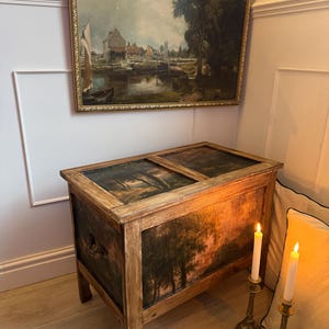Peut inclure: Un coffre en bois ancien avec une peinture de paysage sur le devant et le dessus. Le coffre a un cadre marron clair et des panneaux peints en vert fonc&eacute; et marron. Une peinture de paysage encadr&eacute;e est accroch&eacute;e au-dessus du coffre.