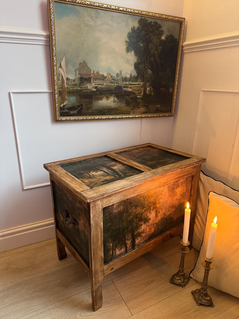 Peut inclure: Un coffre en bois ancien avec une peinture de paysage sur le devant et le dessus. Le coffre a un cadre marron clair et des panneaux peints en vert fonc&eacute; et marron. Une peinture de paysage encadr&eacute;e est accroch&eacute;e au-dessus du coffre.
