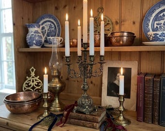 May include: A five-arm brass candelabra holding lit white taper candles, positioned on stacked antique books with velvet ribbons. Flanked by two brass candlesticks with lit candles, a brass oil lamp, copper bowls, and vintage books.