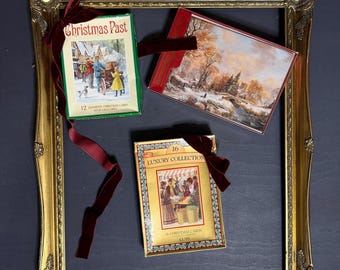 May include: A gold-colored ornate picture frame holds three boxes of Christmas cards. The boxes are tied with burgundy velvet ribbons. The boxes feature vintage illustrations and text, including "Christmas Past" and "Luxury Collection".