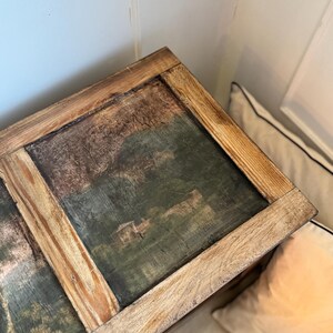 Peut inclure: Un coffre en bois avec une peinture de paysage sur le dessus. La peinture pr&eacute;sente des nuances de vert, de marron et de bleu. Le coffre a un cadre en bois brun clair. Le coffre est plac&eacute; dans un coin &agrave; c&ocirc;t&eacute; d'un mur blanc et d'un oreiller.