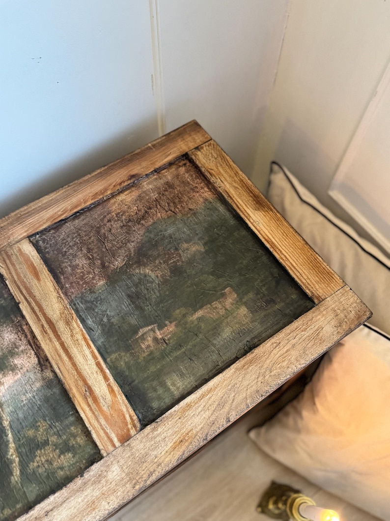 Peut inclure: Un coffre en bois avec une peinture de paysage sur le dessus. La peinture pr&eacute;sente des nuances de vert, de marron et de bleu. Le coffre a un cadre en bois brun clair. Le coffre est plac&eacute; dans un coin &agrave; c&ocirc;t&eacute; d'un mur blanc et d'un oreiller.