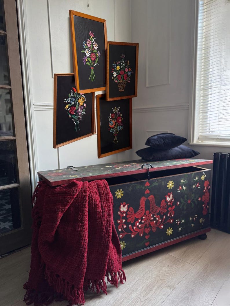 May include: A rectangular wooden storage chest with a black finish, decorated with red bird and floral motifs, featuring a hinged lid. A deep red fringed throw blanket hangs over the side, and four framed floral art pieces hang on the wall above.