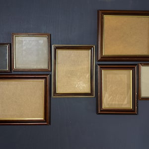 Puede incluir: Una colección de siete marcos de fotos vacíos con bordes de madera marrón oscuro y un fino borde interior dorado. Los marcos varían en tamaño y orientación, incluyendo formas rectangulares y cuadradas, para exhibir obras de arte o fotografías.