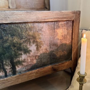 Peut inclure: Gros plan sur un banc ancien en bois avec une peinture de paysage sur le panneau avant. La peinture repr&eacute;sente des arbres et un coucher de soleil. Une bougie allum&eacute;e dans un bougeoir dor&eacute; est au premier plan.