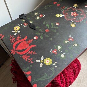 May include: A rectangular black wooden chest lid featuring hand-painted floral motifs in red, yellow, and green. The surface displays a central decorative pattern, smaller scattered flower designs, and a single black metal hinge attached to the top.
