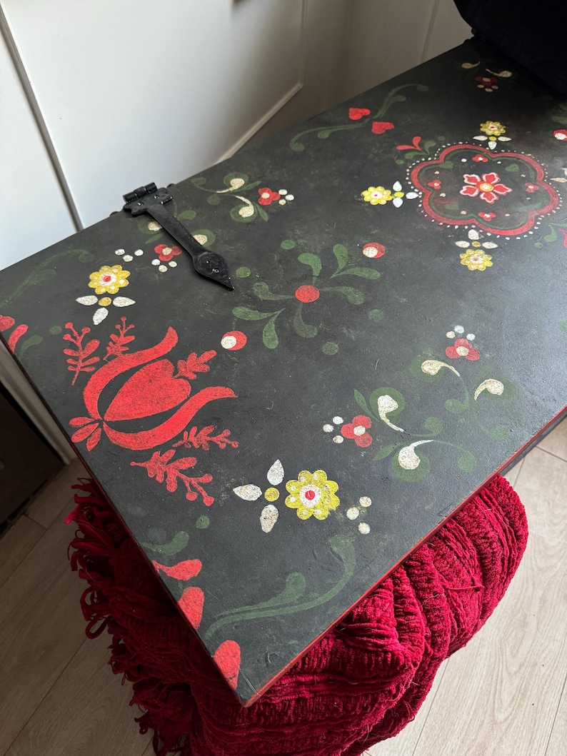 May include: A rectangular black wooden chest lid featuring hand-painted floral motifs in red, yellow, and green. The surface displays a central decorative pattern, smaller scattered flower designs, and a single black metal hinge attached to the top.