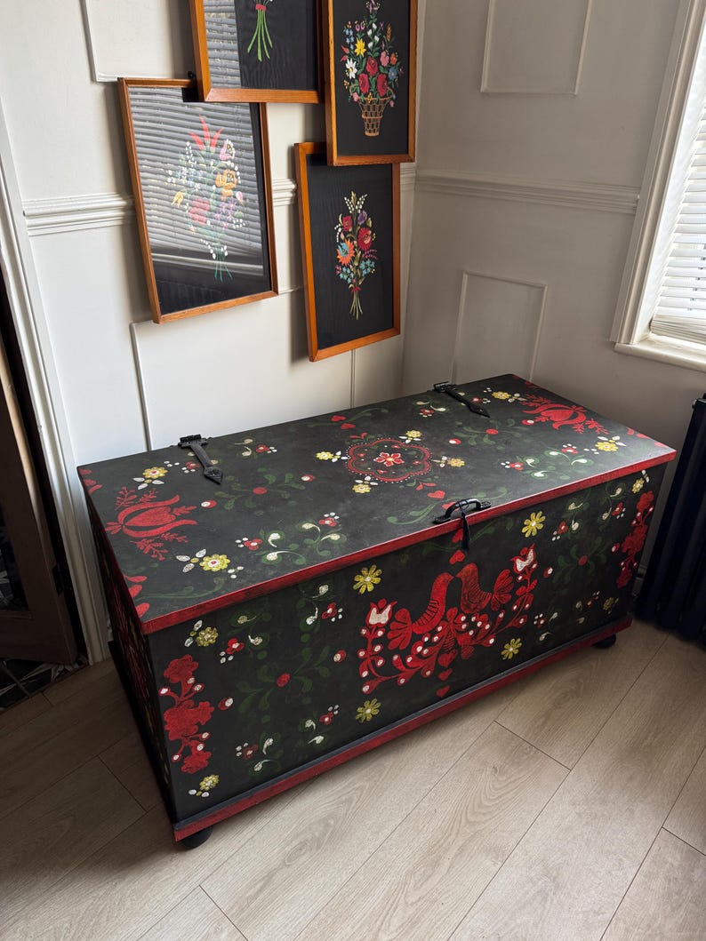 May include: A rectangular wooden storage chest with a black finish, featuring hand-painted floral motifs and bird illustrations in red, yellow, and green. The chest has a hinged lid with black metal hardware and sits on small, dark, rounded feet.