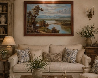 May include: A beige sofa with patterned throw pillows sits before a wooden coffee table holding a ceramic vase, a bowl, and books. Above the sofa hangs a large landscape painting in a dark wood frame, illuminated by a brass picture light.