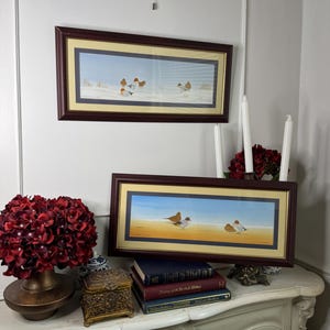 May include: Two framed landscape paintings with bird illustrations, set in brown frames. The paintings depict birds on a beach with a blue and yellow sky. They are displayed above a mantelpiece with books and floral decor.
