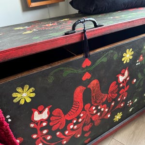 May include: A rectangular wooden storage chest with a black finish and red trim. The front panel features hand-painted folk art motifs including red birds, yellow flowers, and white dots. A black metal handle with a small black tassel is attached.