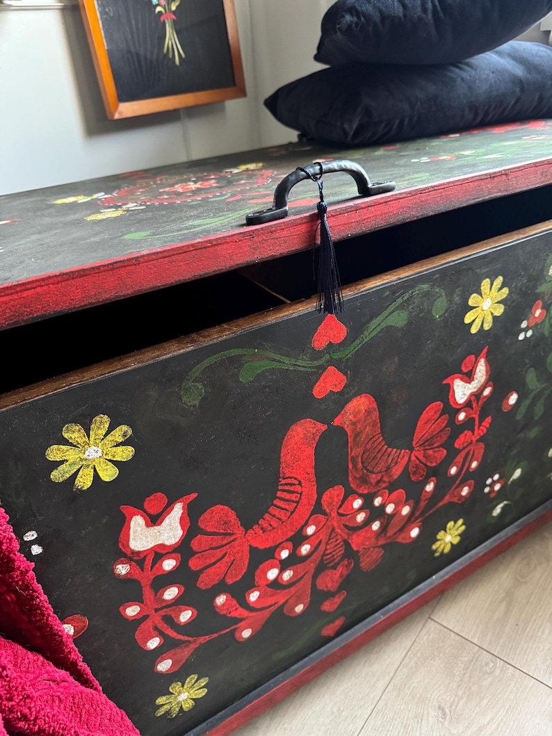 May include: A rectangular wooden storage chest with a black finish and red trim. The front panel features hand-painted folk art motifs including red birds, yellow flowers, and white dots. A black metal handle with a small black tassel is attached.
