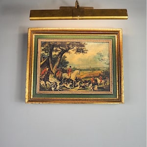 May include: A rectangular framed print depicting a fox hunting scene with riders on horseback and hounds in a field. The frame is gold with a green inner border and a distressed finish. A gold-toned metal picture light is mounted above the frame.