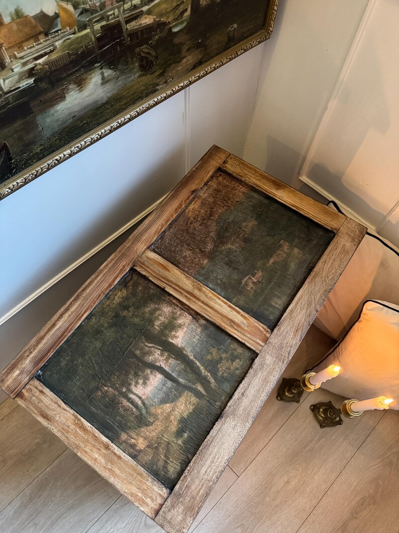Peut inclure: Table basse rectangulaire en bois avec deux peintures de paysage encadr&eacute;es. Les peintures pr&eacute;sentent une sc&egrave;ne de for&ecirc;t aux tons verts et bruns. La table a une finition marron clair vieillie et repose sur un sol en bois clair.