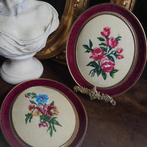 Puede incluir: Dos adornos de pared ovalados con patrones de flores en punto de cruz sobre tela crema. Cada pieza está enmarcada en metal dorado con un borde de terciopelo burdeos. Uno muestra rosas rosadas y el otro un ramo multicolor.