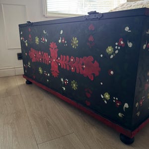 May include: A rectangular black wooden storage chest featuring hand-painted floral motifs in red, white, and green. The chest has a hinged lid, a red base trim, and sits on four small black rounded feet. The front panel displays symmetrical red floral patterns.