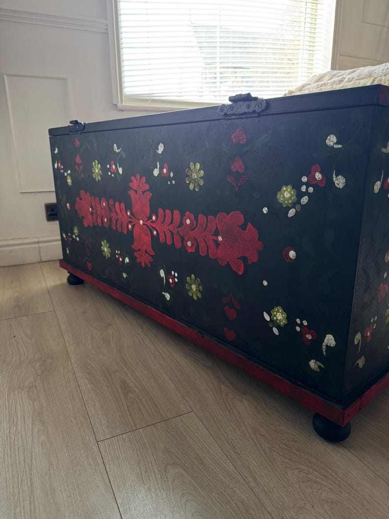 May include: A rectangular black wooden storage chest featuring hand-painted floral motifs in red, white, and green. The chest has a hinged lid, a red base trim, and sits on four small black rounded feet. The front panel displays symmetrical red floral patterns.