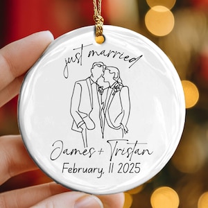 Peut inclure: Ornement rond en céramique blanche avec une ficelle dorée. Il présente un dessin au trait noir d'un couple s'embrassant. Les mots "just married" sont au-dessus du couple, avec les noms et la date "11 février 2025" en dessous.