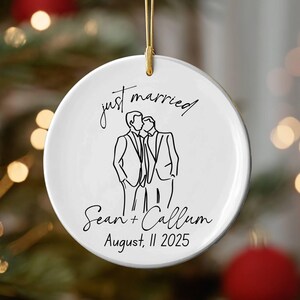 Peut inclure: Ornement de Noël rond en céramique blanche avec une ficelle dorée. Il présente un dessin au trait noir de deux hommes s'embrassant, avec les mots "just married" au-dessus et des noms et une date en dessous.