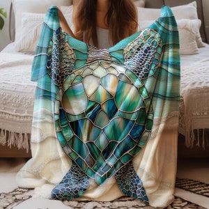 May include: A soft, plush blanket featuring a stained glass style sea turtle design in shades of blue, green, and gold. The blanket is folded in half and held by a person sitting on a bed.