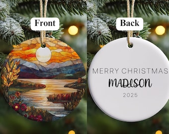 May include: A round Christmas ornament with a landscape design on the front and text on the back. The front features a sunset scene with a lake, mountains, and a full moon. The back says "MERRY CHRISTMAS" and "MADISON 2025".