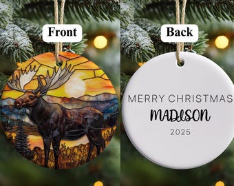 Könnte beinhalten: Runder Weihnachtsschmuck mit Elch-Design. Die Vorderseite zeigt einen Elch in einer Landschaft mit Sonnenuntergang. Auf der Rückseite steht "MERRY CHRISTMAS MADISON 2025". Der Schmuck hängt an einer natürlichen Schnur.