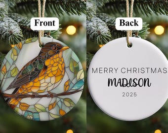 May include: A round Christmas ornament with a stained-glass style bird design on the front. The bird has brown, yellow, and green feathers, perched on a branch. The back of the ornament is white with the text "MERRY CHRISTMAS MADISON 2025".