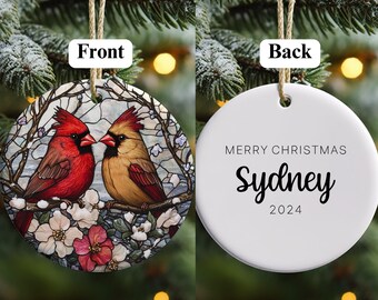 May include: A round ceramic Christmas ornament with a stained glass design of two red cardinals perched on a branch with white blossoms. The back of the ornament is white with the text "Merry Christmas Sydney 2024".