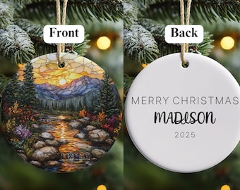 May include: Round Christmas ornament with a landscape scene of a river, mountains, and sunset. The back of the ornament is white with the text "MERRY CHRISTMAS MADISON 2025". The ornament is hanging from a natural twine.