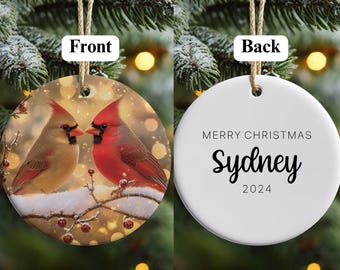 May include: A round Christmas ornament with a front image of two cardinals perched on a snow-covered branch. The back of the ornament is white with the text "MERRY CHRISTMAS Sydney 2024" in black. The ornament hangs from a natural twine.