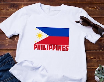 May include: White t-shirt featuring the flag of the Philippines, with blue, red, and yellow colors, and the word "PHILIPPINES" in red. The shirt is paired with blue jeans and sunglasses on a wooden surface.