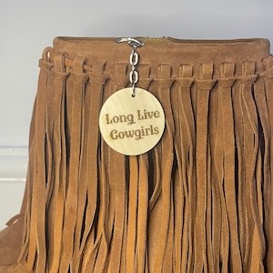 May include: Brown suede fringe boots with a wooden heel. A wooden tag with the text "Long Live Cowgirls" is attached to the boot.