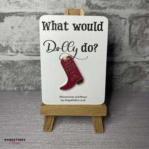 May include: A magenta enamel pin shaped like a cowboy boot with gold trim and the text What would Dolly do? displayed on a white rectangular card. The card features the text What would Dolly do? and is held on a small wooden easel.