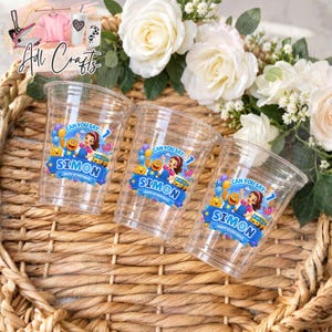 May include: Three clear plastic party cups featuring a colorful graphic of a cartoon character, a yellow school bus, and blue balloons. The text on each cup reads "CAN YOU SAY... 1 SIMON". The cups are arranged on a woven surface with white flowers.