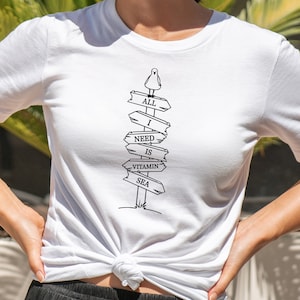 Puede incluir: Camiseta blanca con un dise&ntilde;o gr&aacute;fico en blanco y negro de un poste de se&ntilde;alizaci&oacute;n con las palabras "ALL I NEED IS VITAMIN SEA". Un peque&ntilde;o p&aacute;jaro est&aacute; posado en la parte superior. La camiseta est&aacute; anudada a la cintura.