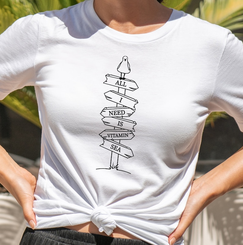 Puede incluir: Camiseta blanca con un dise&ntilde;o gr&aacute;fico en blanco y negro de un poste de se&ntilde;alizaci&oacute;n con las palabras "ALL I NEED IS VITAMIN SEA". Un peque&ntilde;o p&aacute;jaro est&aacute; posado en la parte superior. La camiseta est&aacute; anudada a la cintura.