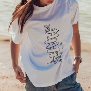 Puede incluir: Camiseta blanca con una ilustraci&oacute;n en blanco y negro de un poste de se&ntilde;alizaci&oacute;n con flechas, un b&uacute;ho, una mariposa y un toc&oacute;n de &aacute;rbol. El dise&ntilde;o est&aacute; centrado en la parte delantera de la camiseta.