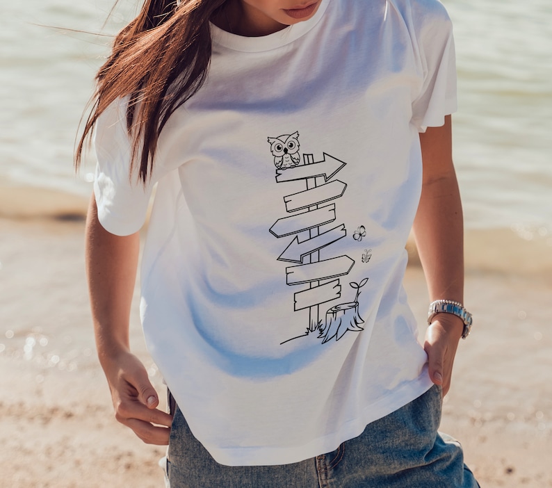 Puede incluir: Camiseta blanca con una ilustraci&oacute;n en blanco y negro de un poste de se&ntilde;alizaci&oacute;n con flechas, un b&uacute;ho, una mariposa y un toc&oacute;n de &aacute;rbol. El dise&ntilde;o est&aacute; centrado en la parte delantera de la camiseta.