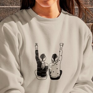 K&ouml;nnte beinhalten: Ein hellbeiger Pullover mit einer schwarzen Grafik von zwei Personen, die sich an den H&auml;nden halten und l&auml;cheln. Die Grafik ist mittig auf dem Pullover platziert.
