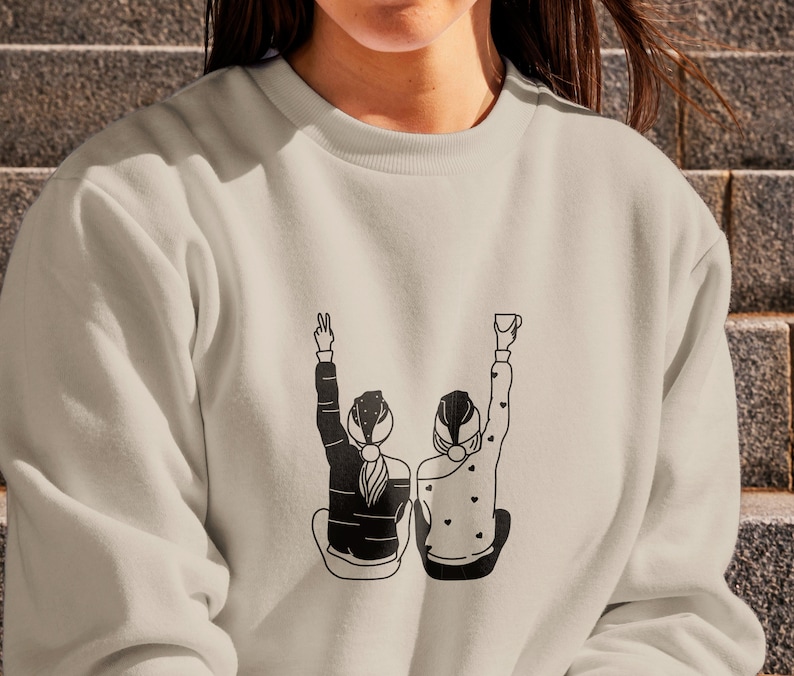 K&ouml;nnte beinhalten: Ein hellbeiger Pullover mit einer schwarzen Grafik von zwei Personen, die sich an den H&auml;nden halten und l&auml;cheln. Die Grafik ist mittig auf dem Pullover platziert.
