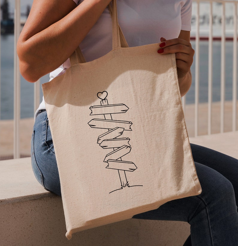 Puede incluir: Bolso tote de lona beige con un dibujo lineal negro de un poste de se&ntilde;alizaci&oacute;n con m&uacute;ltiples flechas. El poste de se&ntilde;alizaci&oacute;n tiene un coraz&oacute;n en la parte superior. El bolso lo sostiene una persona que lleva una camisa blanca y vaqueros azules.