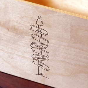 Puede incluir: Letrero de madera con el texto "ALL I NEED IS VITAMIN SEA" grabado en la madera. Un peque&ntilde;o p&aacute;jaro est&aacute; posado en la parte superior del poste indicador. El letrero es de madera clara.