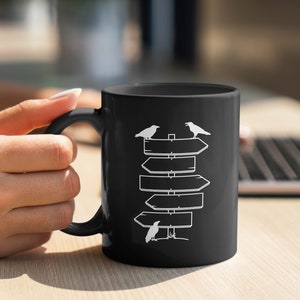 Puede incluir: Taza de cer&aacute;mica negra con un dise&ntilde;o blanco de se&ntilde;ales direccionales y tres cuervos. La taza tiene un asa curvada. El dise&ntilde;o es simple y moderno, adecuado para t&eacute; o caf&eacute;. La taza est&aacute; sujeta por una persona.