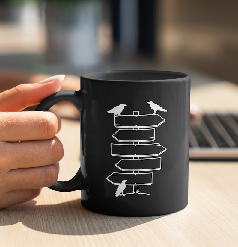 Puede incluir: Taza de cer&aacute;mica negra con un dise&ntilde;o blanco de se&ntilde;ales direccionales y tres cuervos. La taza tiene un asa curvada. El dise&ntilde;o es simple y moderno, adecuado para t&eacute; o caf&eacute;. La taza est&aacute; sujeta por una persona.