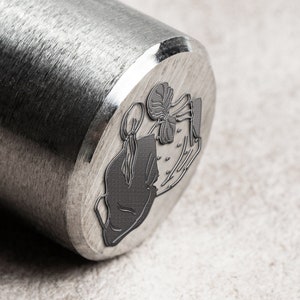 K&ouml;nnte beinhalten: Ein Stempel aus silbernem Metall mit einem Design aus zwei Figuren, die sich die H&auml;nde halten. Die Figuren sind stilisiert und haben ein schlichtes, minimalistisches Design.
