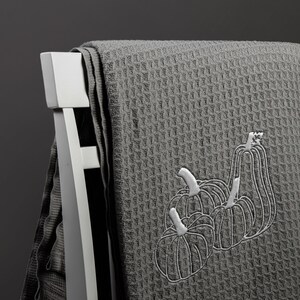 Puede incluir: Una toalla de cocina de tejido de waffle gris con un dise&ntilde;o bordado de tres calabazas blancas.