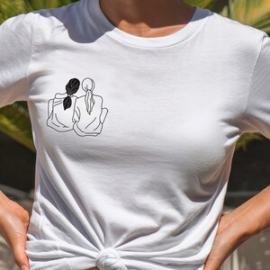 K&ouml;nnte beinhalten: Wei&szlig;es T-Shirt mit einer schwarzen Linienzeichnung von zwei Frauen, die R&uuml;cken an R&uuml;cken sitzen und ihre Arme umeinander gelegt haben.