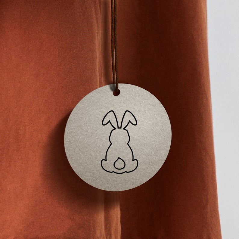 May include: A brown paper tag with a black outline of a rabbit on a brown fabric background.